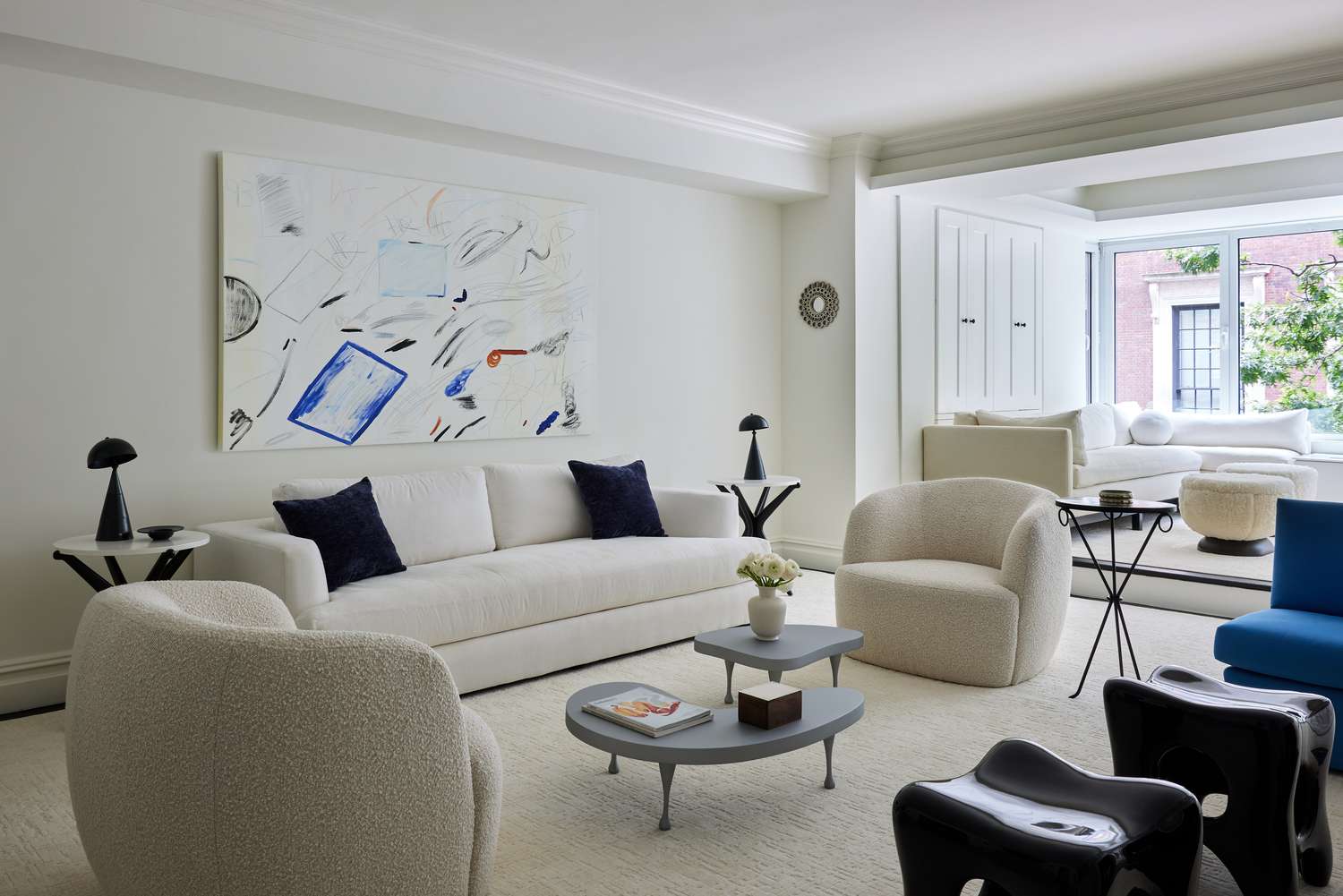 Minimalist living room by Julia Baum Interiors: white textured sectional sofa, cream bouclé chairs, black and silver side tables, blue pillows, and Cy Twombly-inspired artwork. Large window with tree views, neutral rug, and natural light create a calm, stylish space.