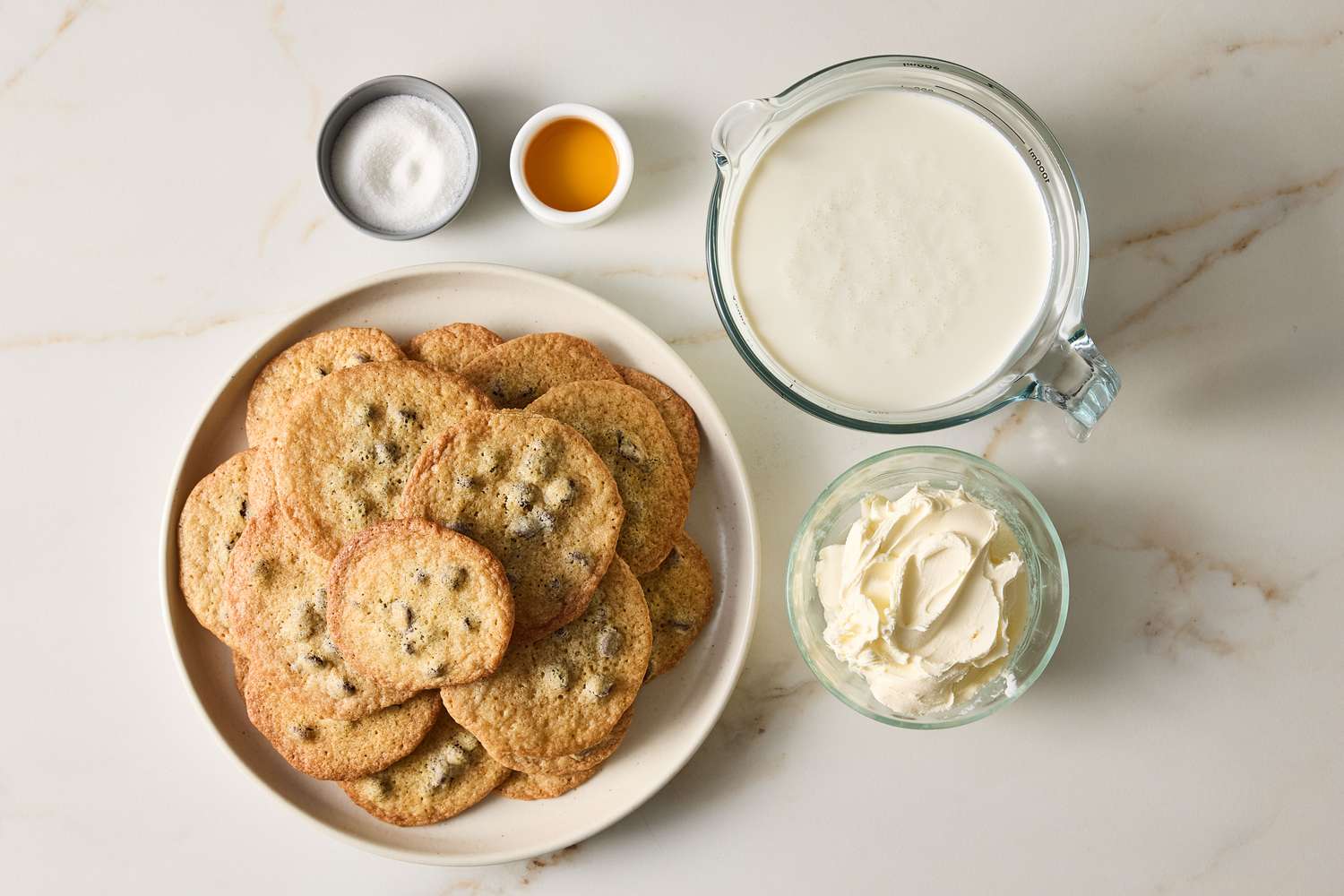 Ingredients for a chocolate chip cookie icebox cake arranged on a surface including cookies cream and small dishes of vanilla and sugar