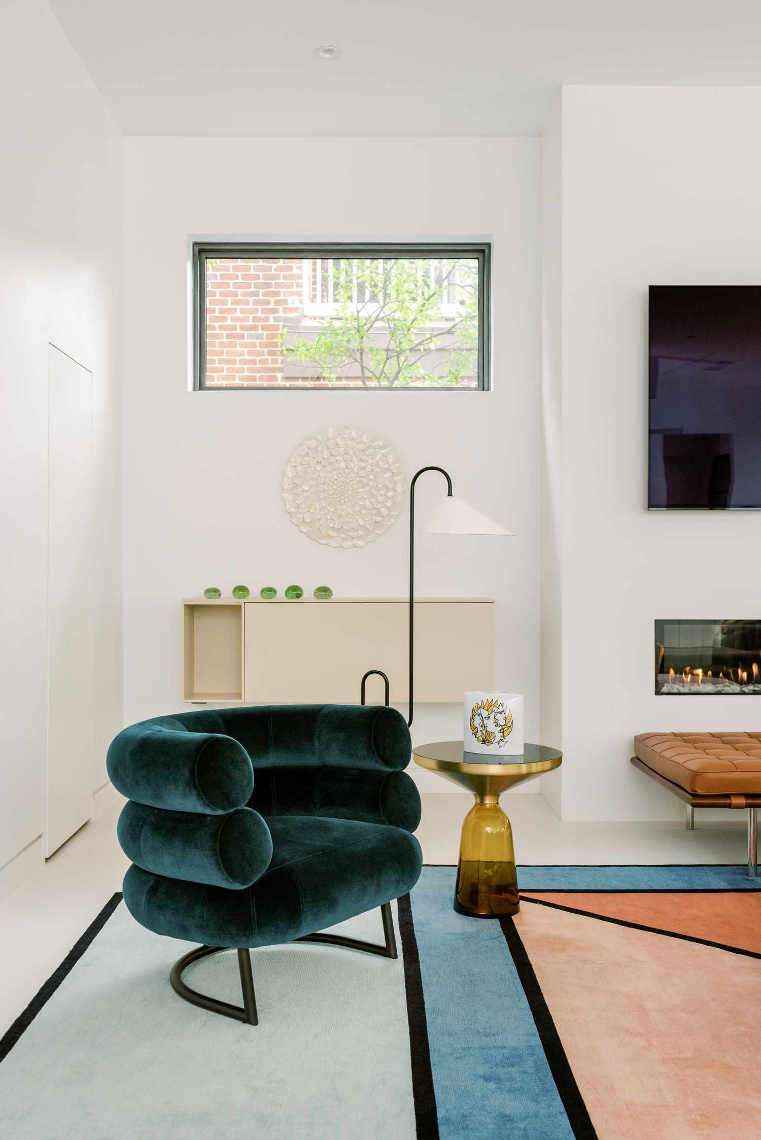 Minimalist living room by Lambrakos Studio: teal velvet armchair with curved design, brass side table, geometric rug in blue and pink, modern fireplace, and decorative wall art. Large window, beige shelving, and natural light create a calm, stylish space.