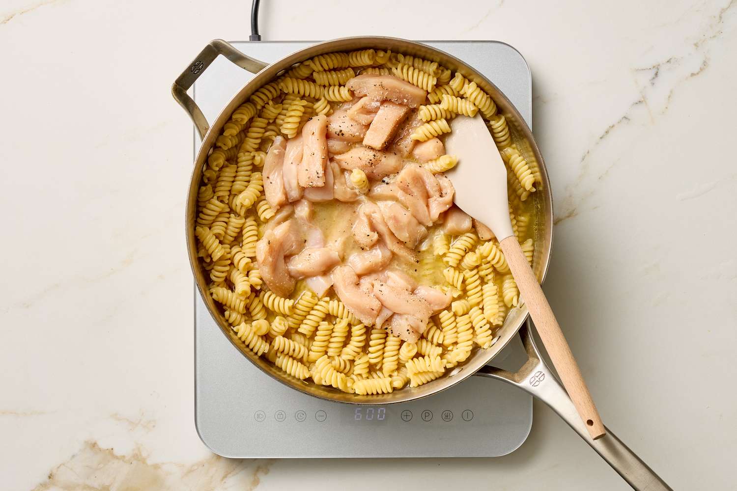 Cooked pasta and sliced chicken in a pan with a spatula on a stovetop