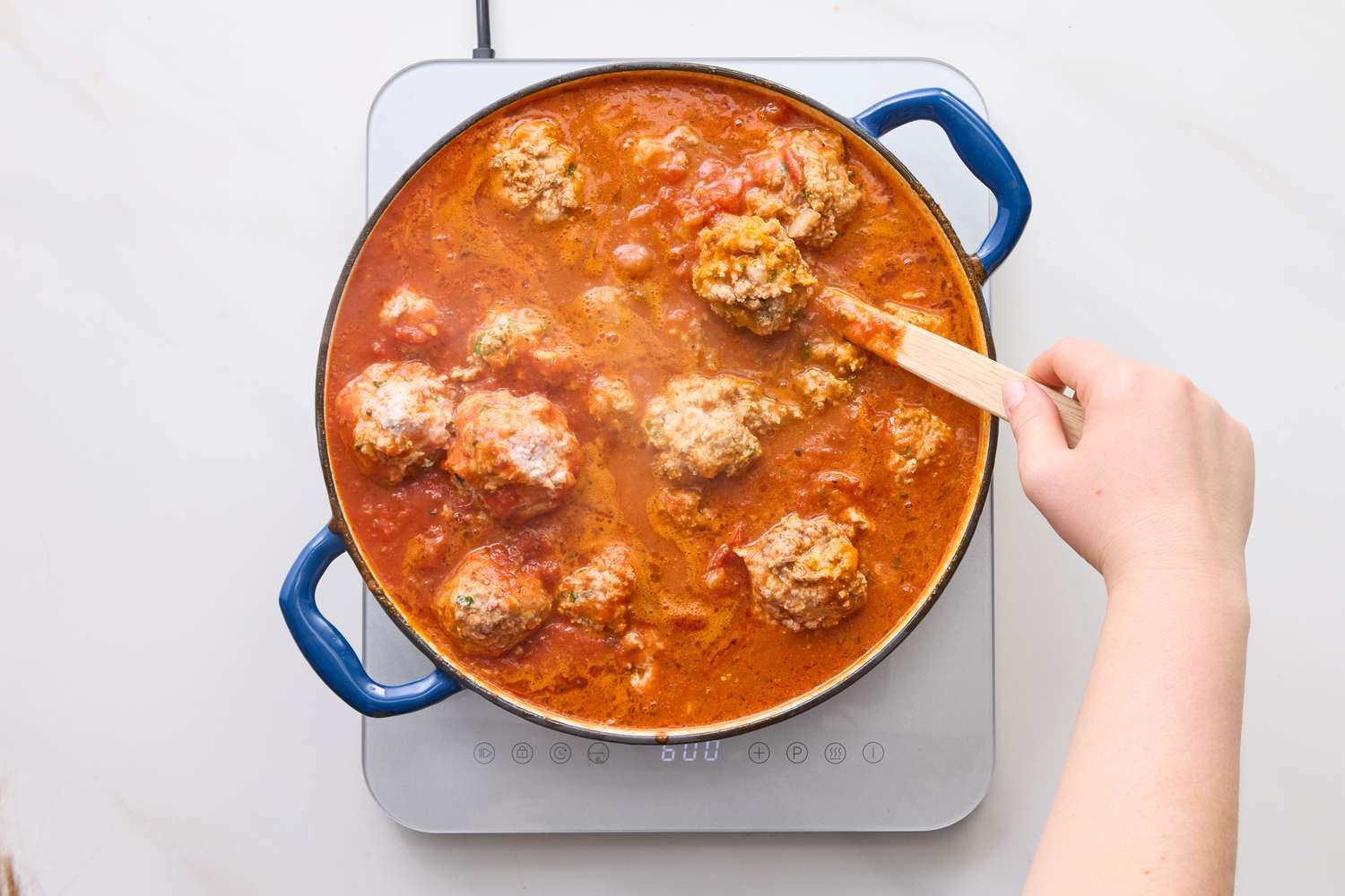 Meatballs cooking in a tomatobased sauce in a pot with a hand holding a spoon
