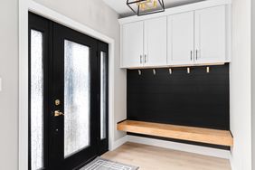 An entryway with a black door white cabinets above a black panel wall and a wooden bench with hooks underneath