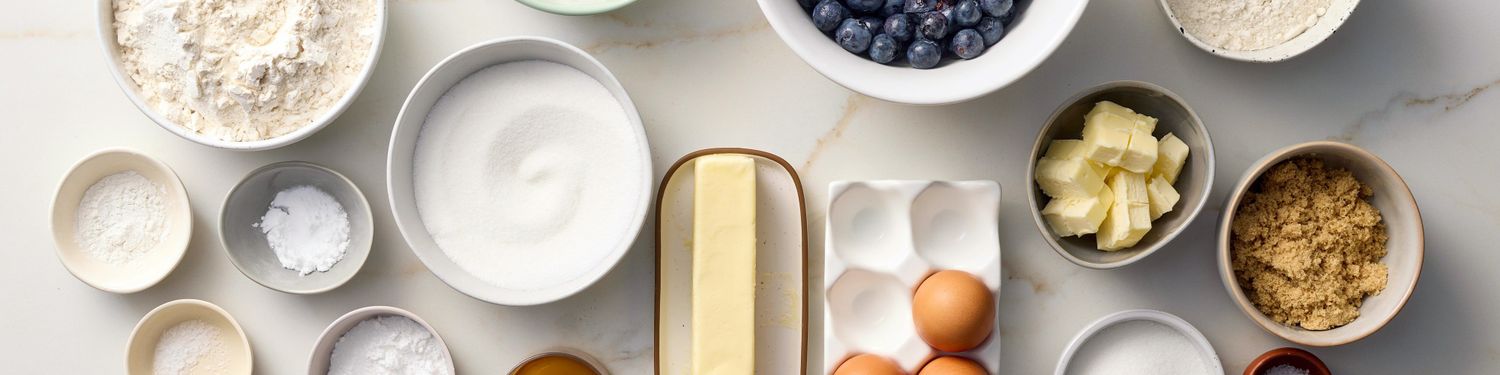 Ingredients for baking laid out on a surface including flour sugar butter eggs and blueberries