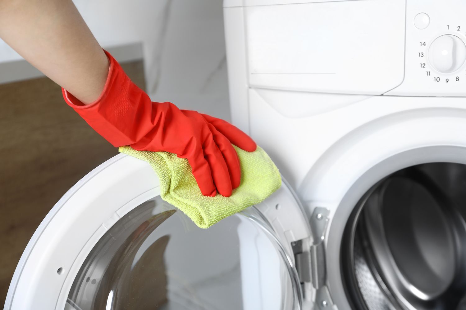 A gloved hand wiping a washing machine door with a yellow cloth