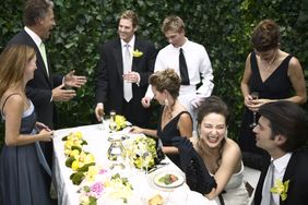 A wedding scene with a group of people including David Corenswet gathered around a table talking and laughing