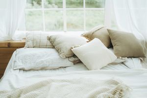 Heap of cushions and pillows on a bed with blanket and plaid against a window with curtains in ground floor room.