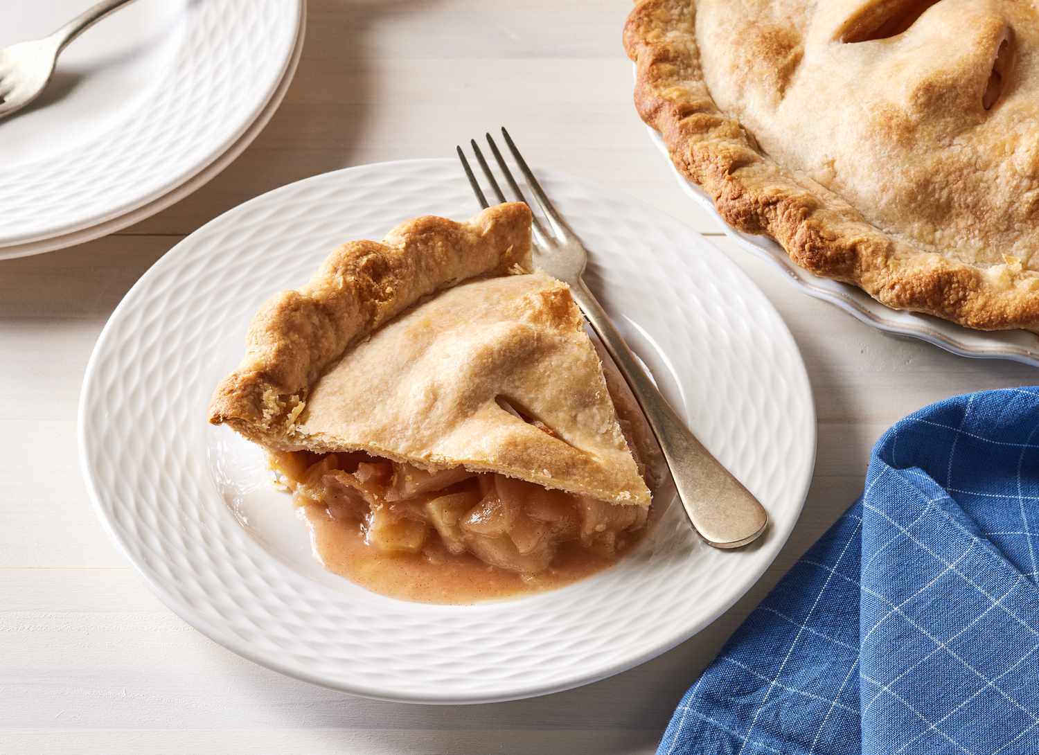 slice of apple pie on white plate with fork, whole pie and blue napkin nearby
