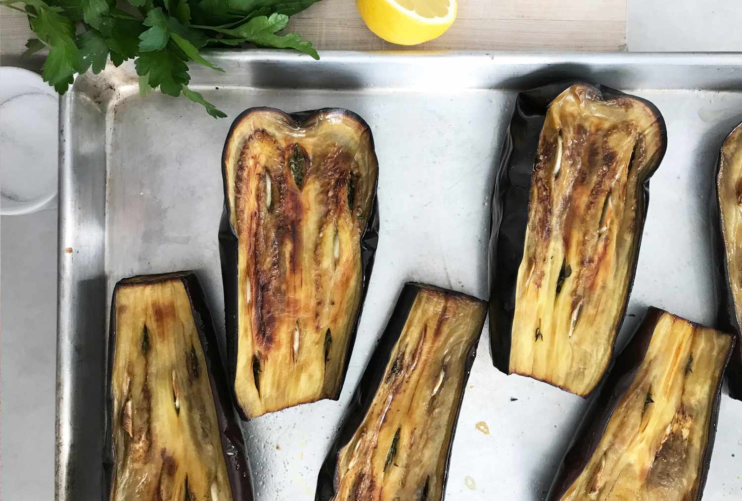 Roasted eggplant halves on a baking sheet with parsley and a lemon wedge nearby