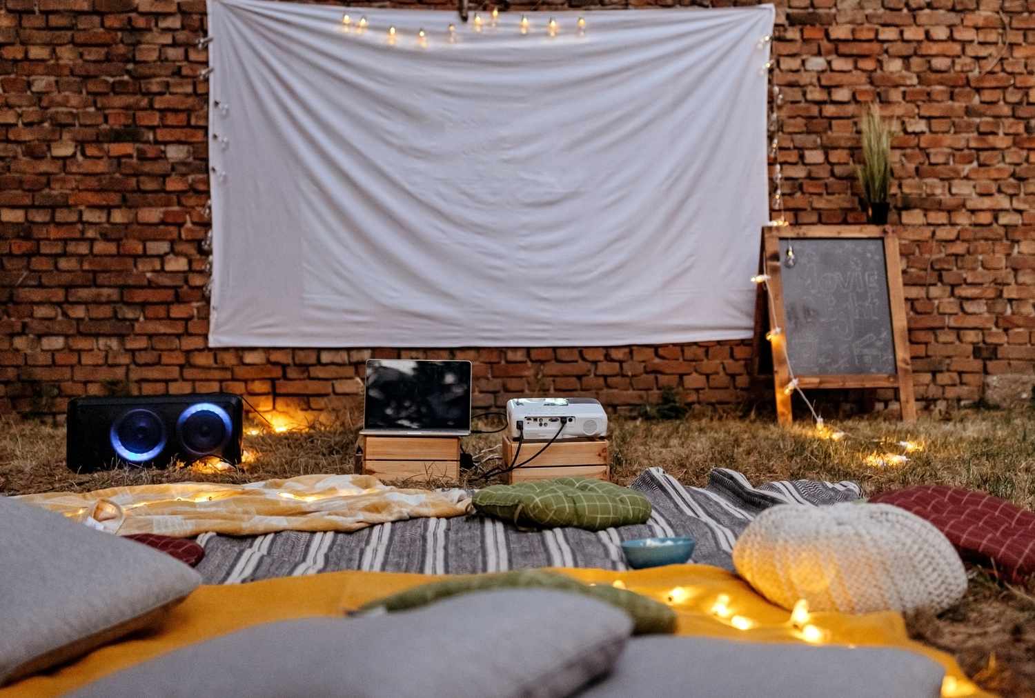 Backyard movie setup with a white screen a projector speakers cushions and lights arranged against a brick wall backdrop
