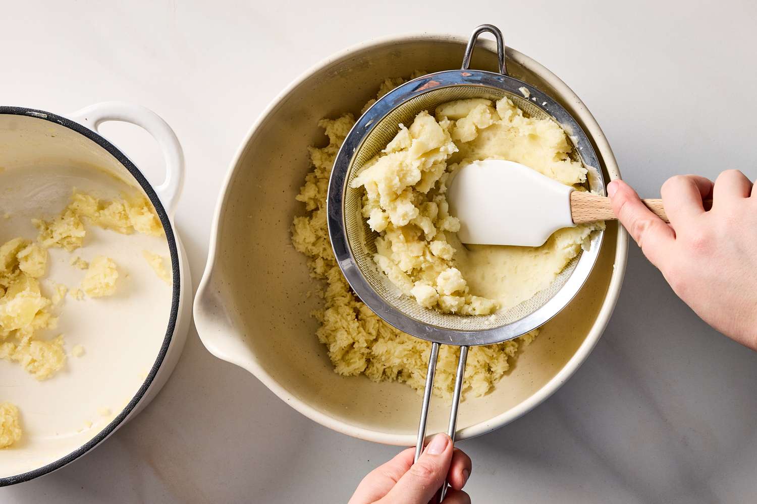 Two hands using a spatula to press cooked potatoes through a sieve into a bowl