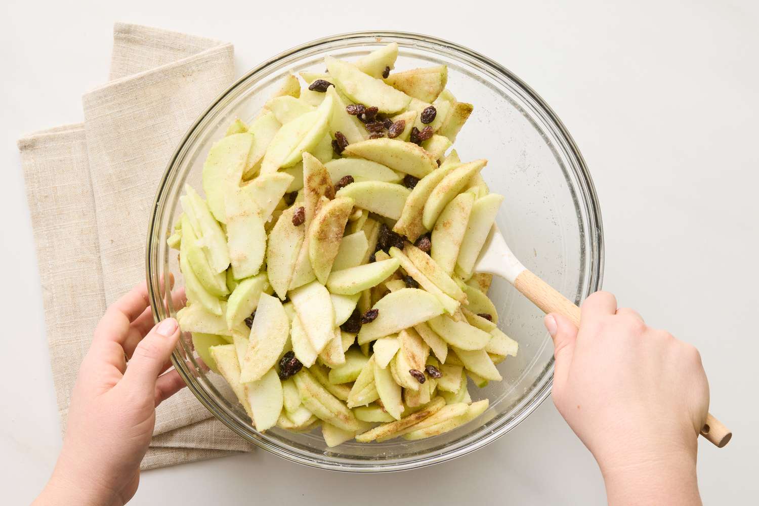 Hands stirring sliced apples and raisins in a glass bowl with a spatula