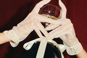 A person wearing lace gloves holds a wine glass over their head decorated with a ribbon tied in a bow