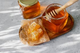 honey in two glass jars, one with lid off and honey dipper, also honeycomb on paper in dappled light
