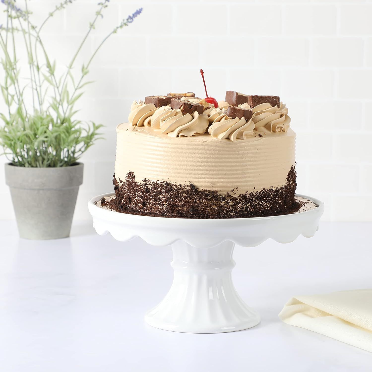 A layered cake with chocolate decoration and a cherry on top displayed on a white pedestal