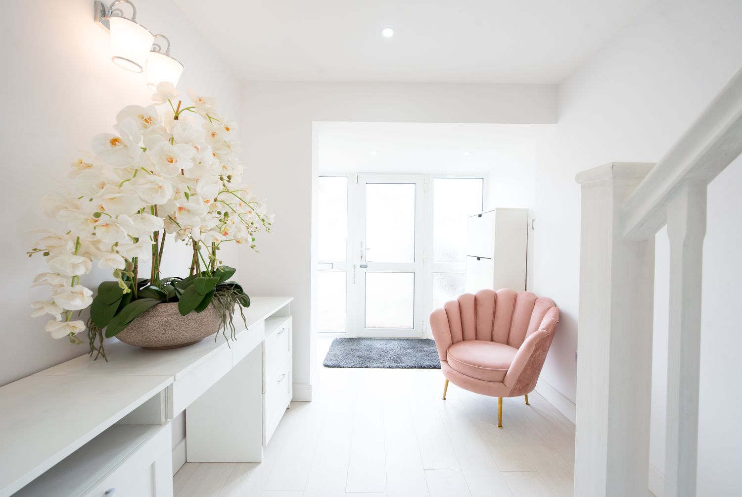 A hallway with a light pink chair and a potted floral arrangement on a cabinet