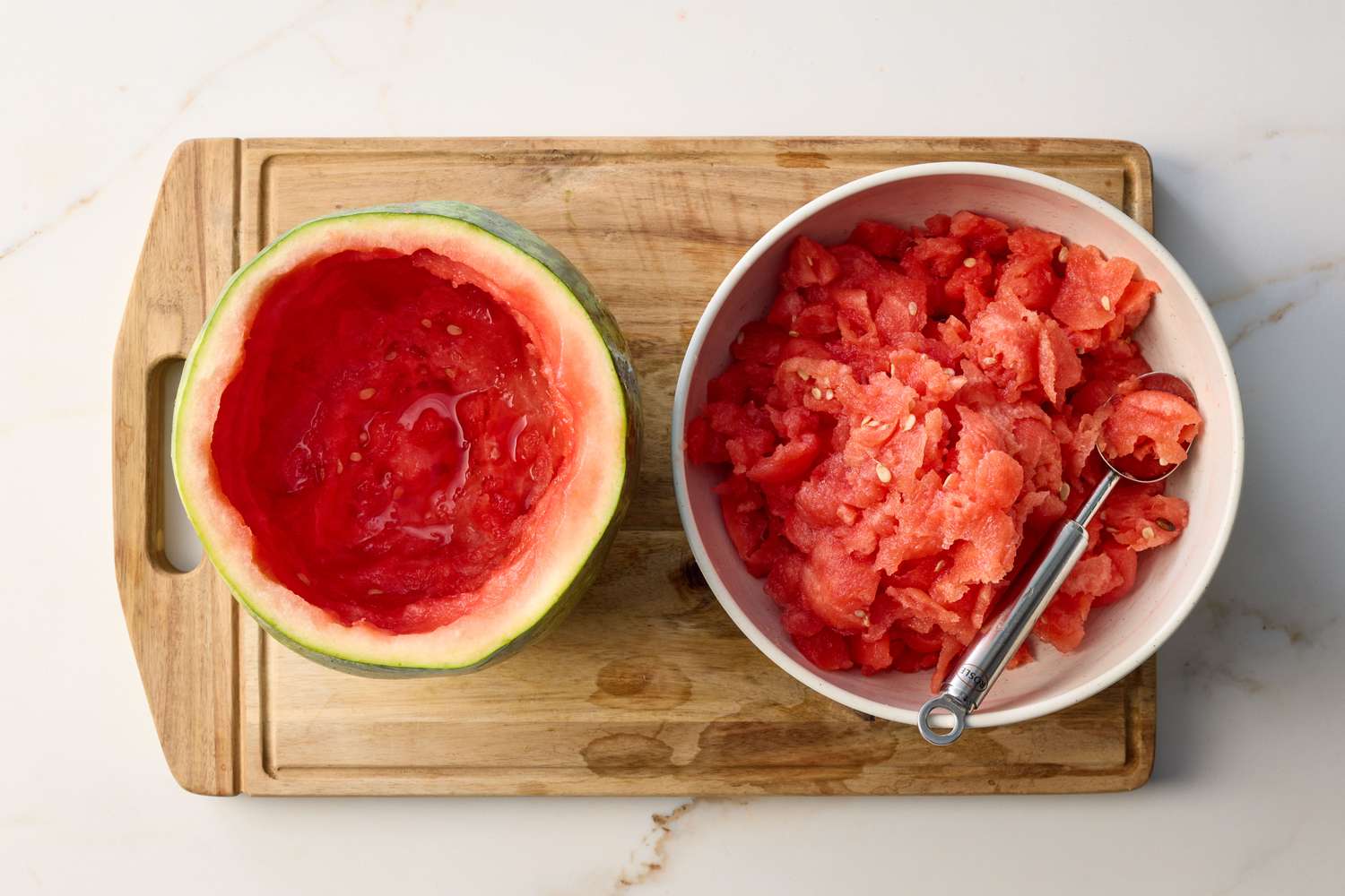 A hollowed-out watermelon on a cutting board next to a bowl filled with scooped watermelon pieces and a spoon